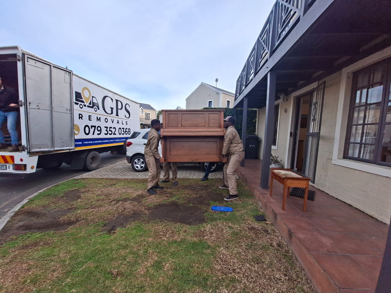 Three GPS Removals movers carrying an upright piano into a house