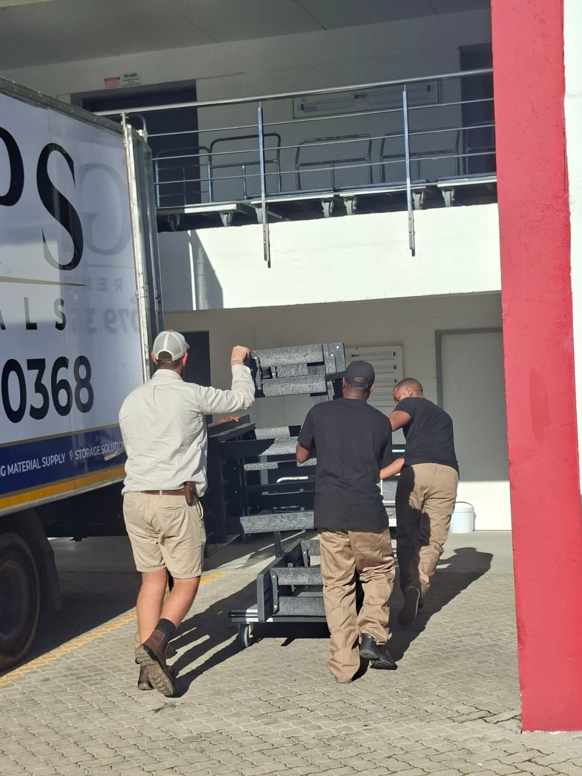 Three movers wheeling a loaded trolley of office furniture towards the truck